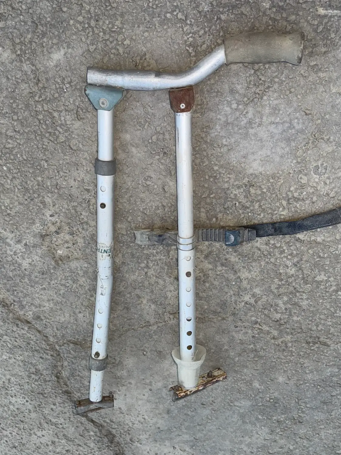 The 2011 hand control laid out on concrete: two silver crutch tubes with rows of adjustment holes, a dark strap, and a curved handle with a rubber grip.