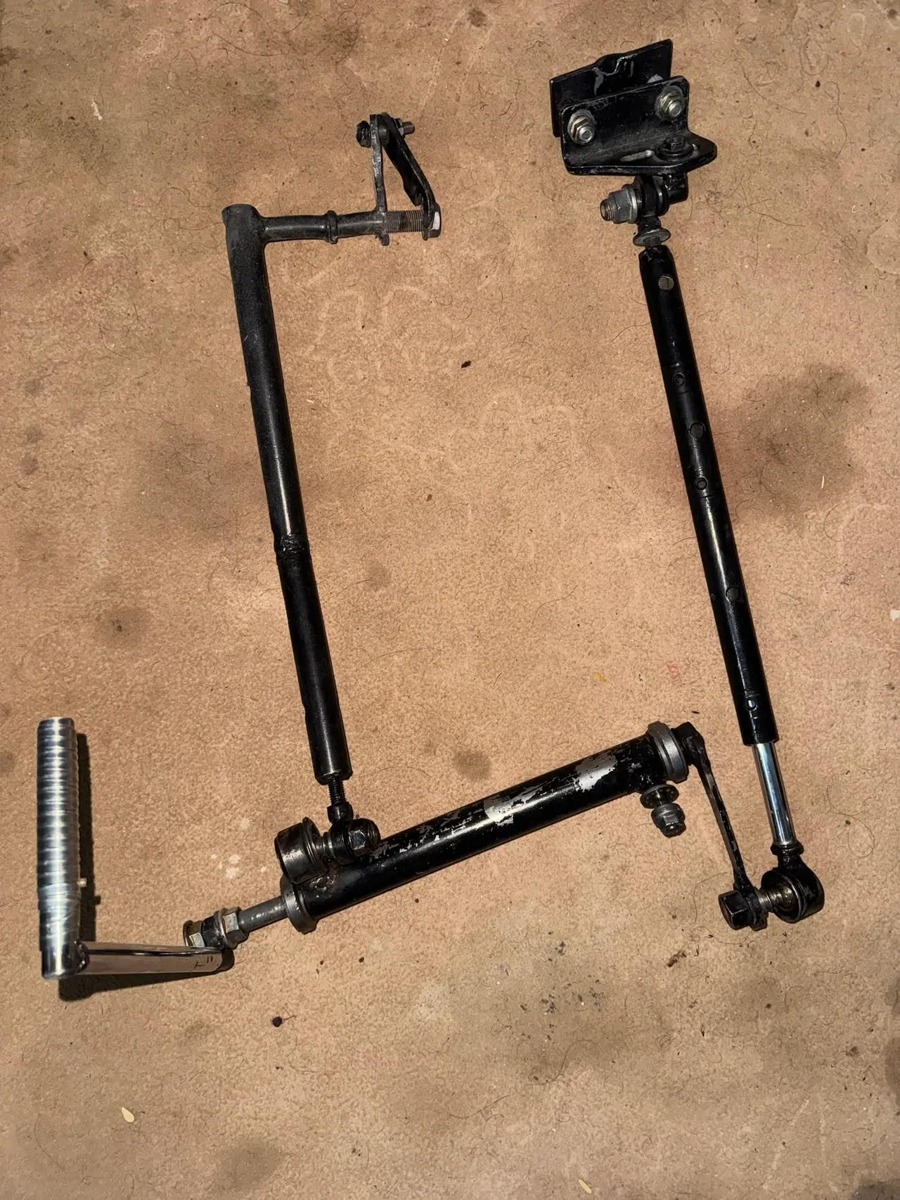 The 2025 hand control: black-painted rods joined by ball joints, with a mounting bracket at one end and a knurled metal grip at the other.