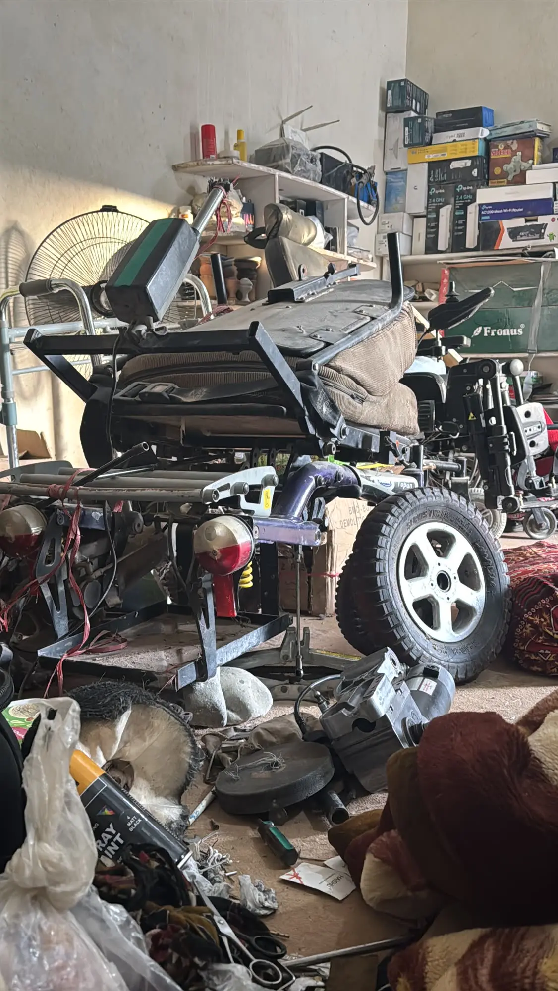 A powered wheelchair part-way through repair in the workshop, its seat frame open, a road wheel and the drive motor set down beside it.