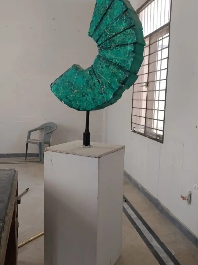 A thesis sculpture: a green segmented arc on a steel post, standing on a plinth in a bare studio.