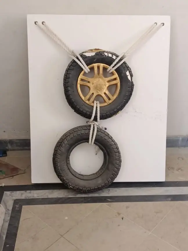 A thesis piece: a wheelchair tyre with a gold-painted hub and a second bare tyre, roped to a white panel.